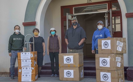 Men and women standing in back of boxes of protective gear used to fight covid-19.