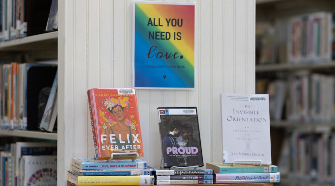 Pride Month book display with stacked books and poster with "All you need is love."