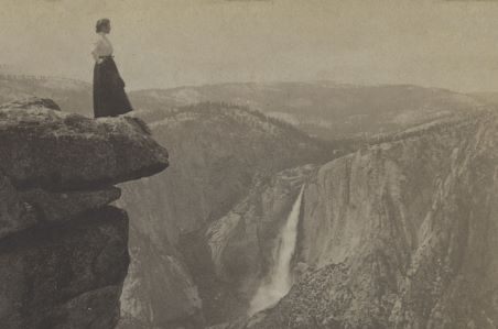 Black and white photo of a woman standing on a high cliff overlooking Yosemite, a waterfall can be seen in the distance. 