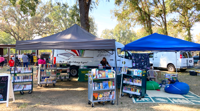 Carts with books and chairs with pop-up kanopies overhead, and outreach van in background at outside event.