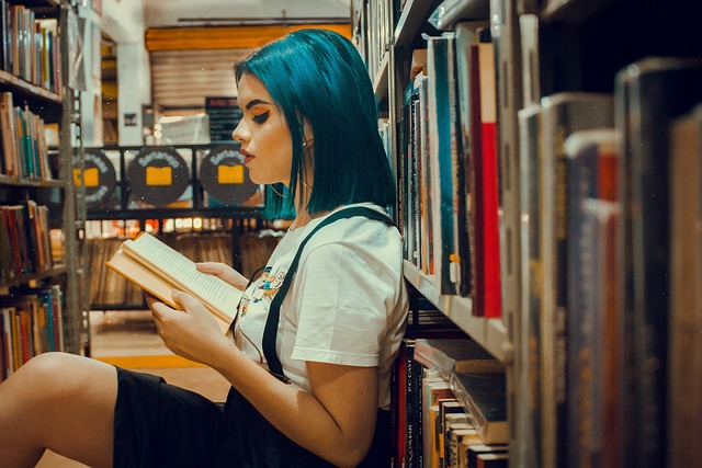 A young woman with blue and black hair sits between two library stacks and reads a book